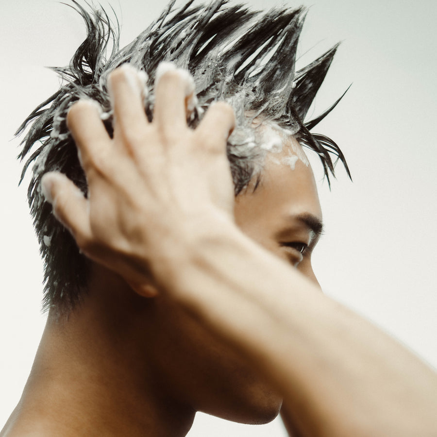 Un jeune homme aux cheveux courts frotte énergiquement le nettoyant/shampoing micellaire sur sa tête, créant une mousse abondante. Ses cheveux courts sont dressés en pics, illustrant l'effet nettoyant et revitalisant du produit.