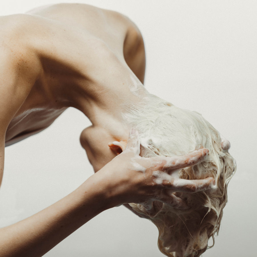 Une jeune femme renversée tête en bas utilise le nettoyant/shampoing micellaire ,pour ses cheveux avec une grande générosité de mousse. Cette photo capture le moment où le produit crée une mousse abondante, illustrant son efficacité pour un nettoyage en profondeur et revitalisant des cheveux.